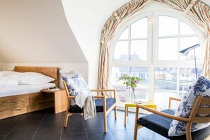 Een slaapkamer met een houten bed, een nachtkastje, een stoel, een kleine tafel met een vaas en een groot raam met gordijnen.
