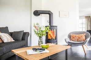 Un salon moderne comprenant un canapé, un fauteuil, une table, un vase avec des fleurs et un poêle à bois.