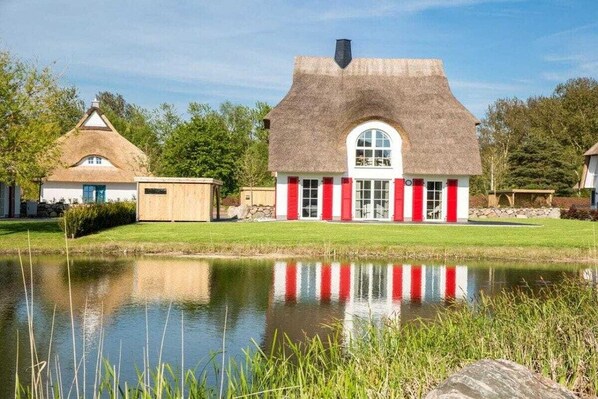 Een huis met rieten dak en rode luiken, gelegen naast een vijver met een heldere spiegeling.