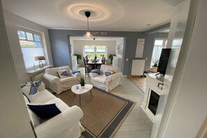 A modern living room with a fireplace, white sofas, a dining table, and a ceiling light fixture. - Holiday apartment with 2 bedrooms (Travemünde)