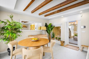 Un comedor con una mesa de madera redonda, sillas color beige y un cuenco con fruta. Hay plantas en macetas y un espejo grande en la pared.