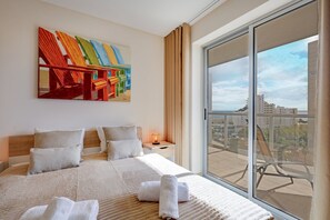 Ein Hotelzimmer mit einem großen Bett, einem Bild an der Wand, einem Nachttisch mit Lampe und einem Balkon mit Blick auf die Stadt.