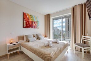 Ein modernes Schlafzimmer mit einem großen Bett, einem Nachttisch mit Lampe, einem Sessel und Blick auf Gebäude durch das Fenster.