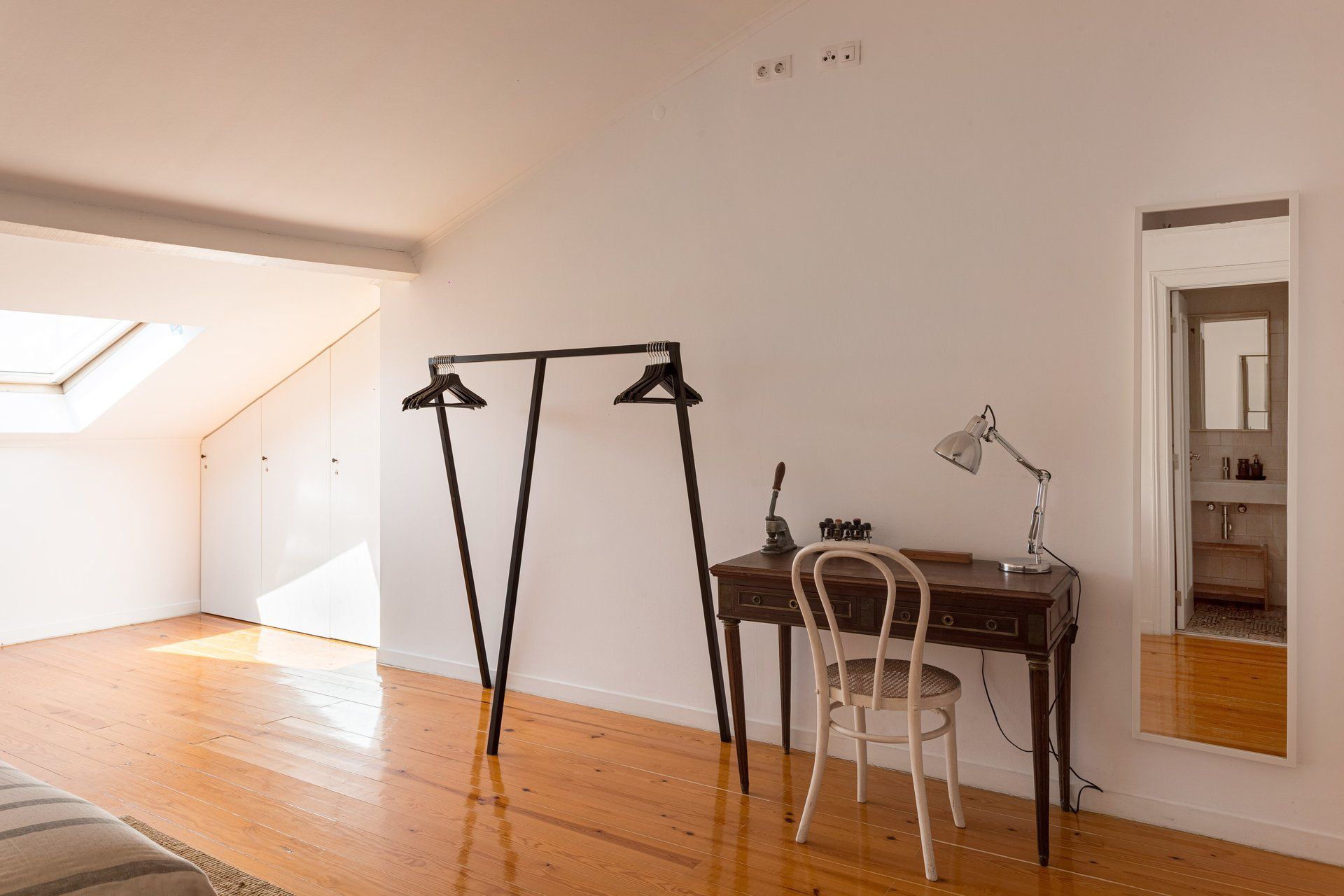 Een kamer met houten vloer, een bureau met stoel, een hangende kledingrek, een bureaulamp en een dakraam.