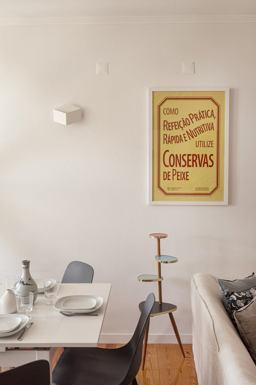 Een eetgedeelte met een witte tafel gedekt voor twee personen, een zwarte stoel, een decoratieve vloerlamp en een ingelijste poster aan de muur.