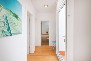 Un pasillo con piso de madera, paredes blancas y una puerta que da a un baño con cabina de ducha azulejada.
