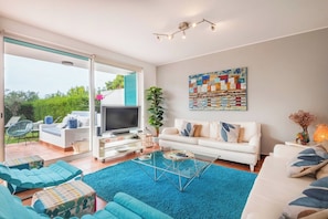 Een moderne woonkamer met een televisie, een witte bank, blauwe stoelen en een groot raam.