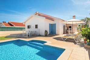 Ein Schwimmbecken mit einem weißen Gebäude, einer überdachten Liegefläche inklusive weißem Sonnenschirm und einem eingezäunten Garten.