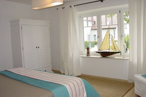 Ein Schlafzimmer mit Bett, Kleiderschrank, einem Fenster mit einem Modell eines Segelboots und Blick auf ein Haus.