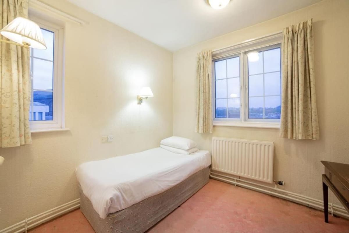 Ein Einzelbett in einem Zimmer mit Fenster, Heizkörper und Lampe.
