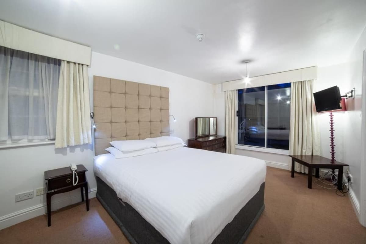 Ein Hotelzimmer mit einem großen Bett, einem Nachttisch, einem Fernseher und einem Fenster mit Vorhängen.