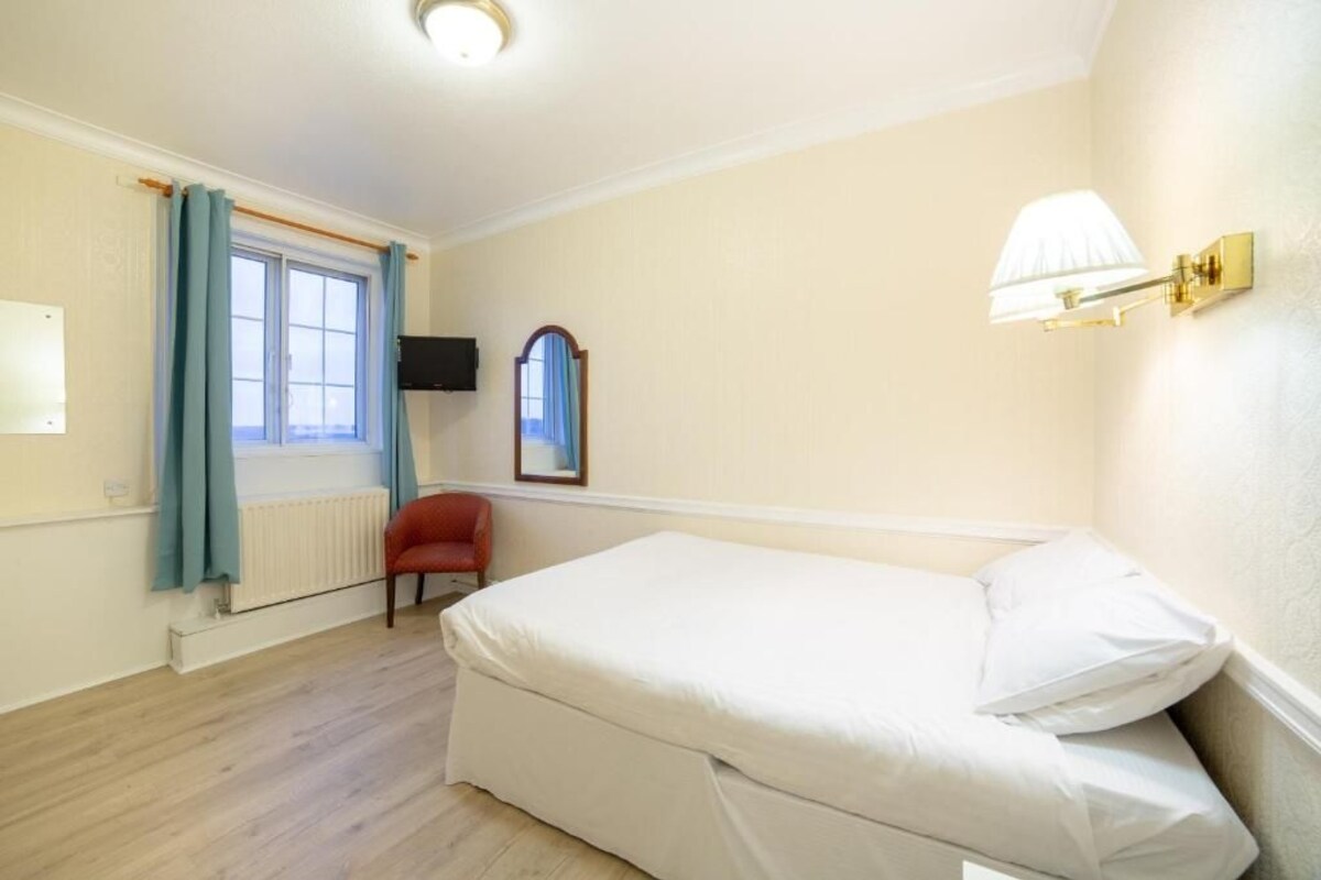 Ein Schlafzimmer mit Bett, Sessel, Fenster mit Vorhängen, Heizkörper, Wand-Fernseher und Wandlampe.