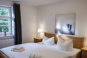 A hotel room with a bed, a nightstand, a lamp, a window with curtains, and a painting on the wall. - House on the Bodden shore (Born a. Darß)