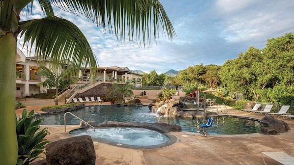 Área de piscina de resort com várias piscinas, incluindo uma banheira de hidromassagem, rodeada por palmeiras e espreguiçadeiras.