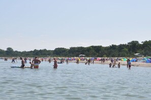 En fullsatt strand med folk i vattnet och på sanden, tält och träd i bakgrunden.