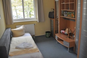 A bedroom with a bed, a desk, a radiator, a window with curtains, and a bookshelf with books and a lamp. - Holiday apartment with 2 bedrooms (Ostseebad Boltenhagen)