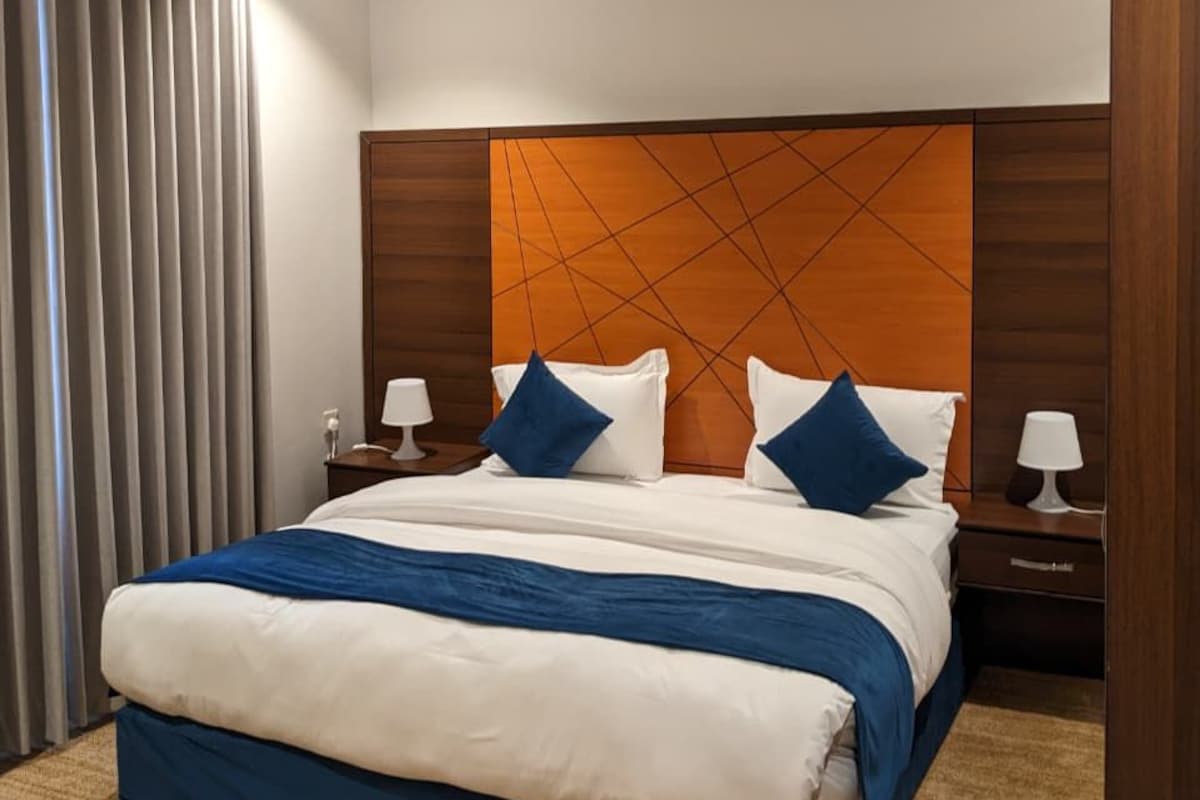A hotel room with a large bed, wooden headboard, two bedside tables with lamps, and a view of the city through the window.
