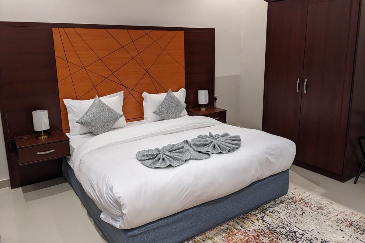 A modern bedroom with a large bed, wooden headboard, nightstands, and a wardrobe.