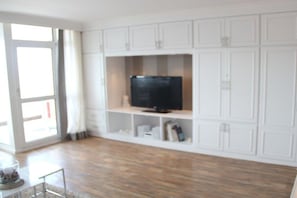 A living room with a television on a white cabinet, a glass coffee table, and a view through a large window. - Holiday apartment in Sylt with 2 bedrooms (Sylt \/ OT Westerland)
