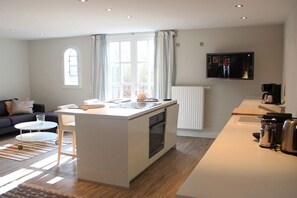 A modern kitchen with a central island, a dining area with a table and chairs, a sofa, and a television mounted on the wall. - Holiday apartment in Sylt with 1 bedroom (Sylt \/ OT Westerland)