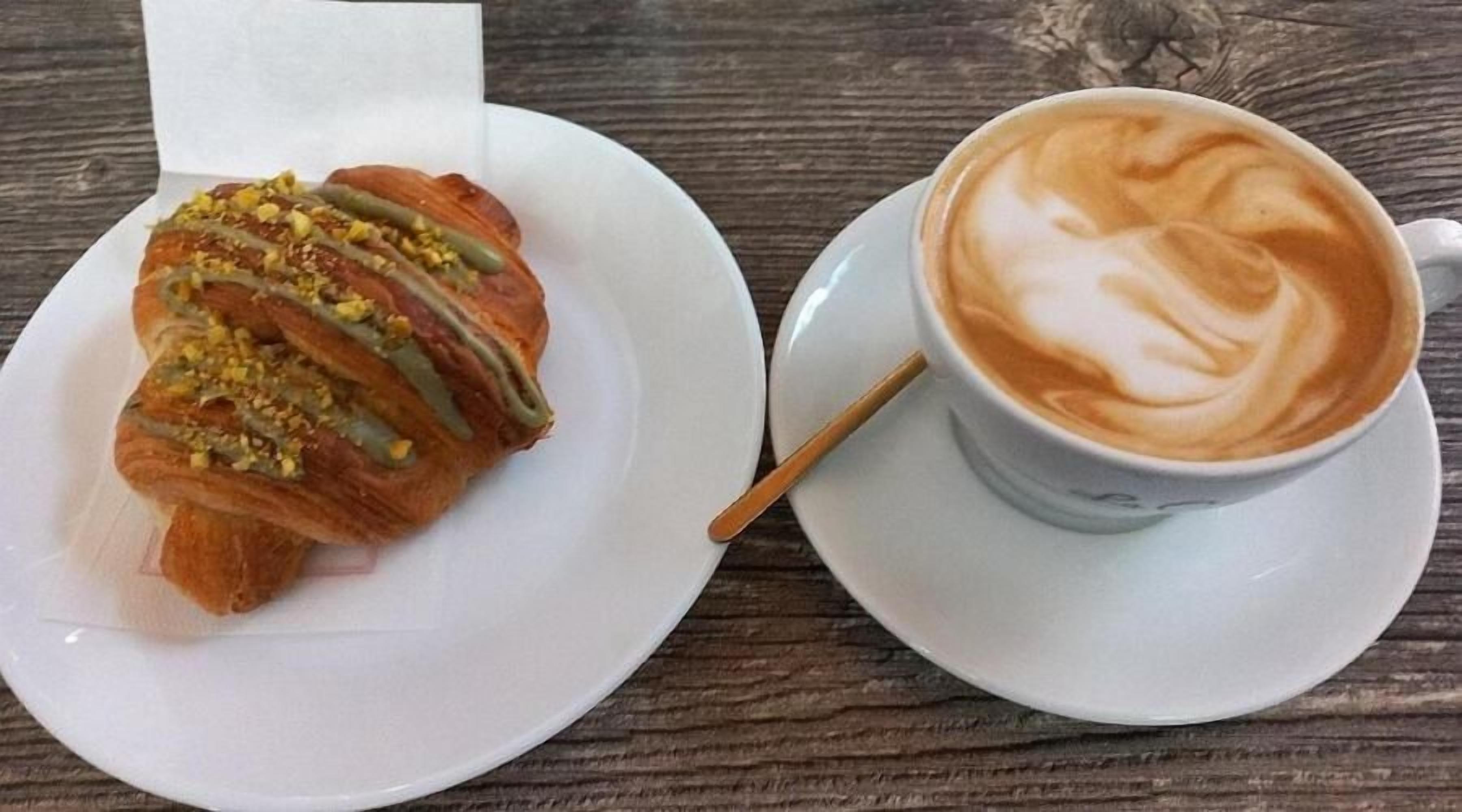 A croissant with a drizzle of chocolate and chopped nuts on a white plate, next to a cup of coffee with latte art on a saucer.
