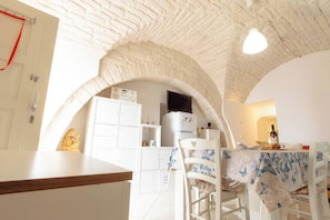 A room with a white arched ceiling, a dining table with chairs, a white cabinet, and a small kitchen area with a refrigerator and a television. - Historic Stone Vault in Noci Incl Breakfast (Noci)