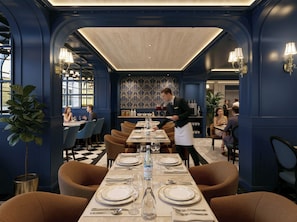 Ein Restaurant mit einem langen Esstisch, der für eine Mahlzeit gedeckt ist, einem Kellner, der sich um Gäste kümmert, und einer gemusterten Wand im Hintergrund.