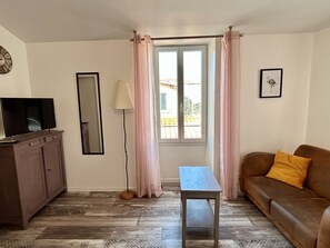 Ein Wohnzimmer mit einer Couch, einem Fernseher auf einem Schrank, einem Tisch, einer Lampe und einem Fenster mit Vorhängen.