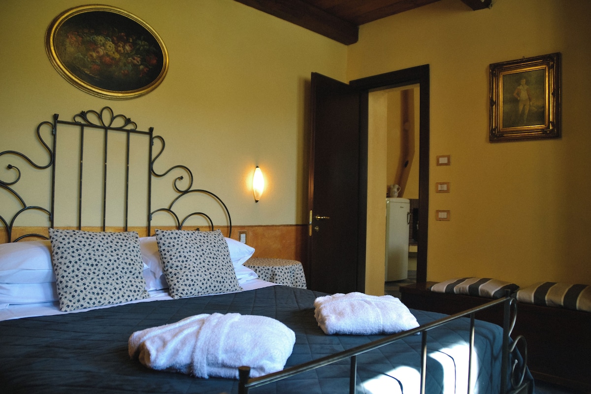 A neatly made bed with patterned pillows and a dark bedspread, a wall-mounted light, a framed picture, and a door leading to another room.