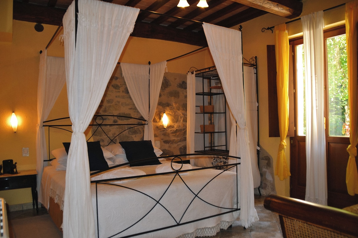 A bed with a metal frame, white bedding, and black pillows. There is a stone wall behind the bed and a wooden ceiling.