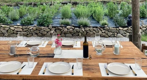 A wooden dining table set for four with white plates, cutlery, and glasses, set against a backdrop of a stone wall and lush greenery. - OASI LIGNANA Apartments (PESCIA)