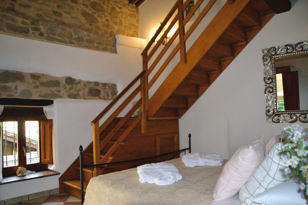 A bedroom with a wooden staircase, a bed with white linens, a stone wall, and a mirror with an ornate frame.