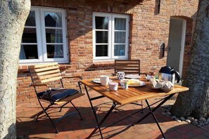 An outdoor dining area with a wooden table and chairs, a brick wall, and a window. - Holiday apartment with 1 bedroom (Alkersum)