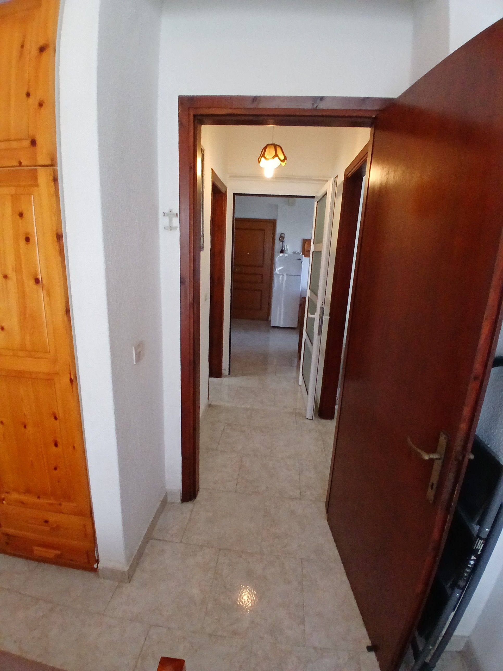 A hallway with a wooden door on the left, a white wall, and a light fixture above a door on the right.