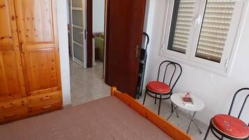 Ein Schlafzimmer mit Bett, Holzschrank, einem kleinen Tisch mit Stühlen und einem Fenster mit Jalousien.