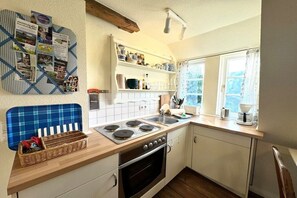 A kitchen with a stove, sink, and window. There is a blue chair and a shelf with dishes. - Old registry office studio apartment (Oldsum)