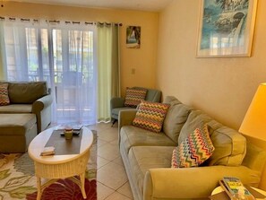 A living room with a sofa, a coffee table, and a chair. There is a window with curtains, a painting on the wall, and a lamp on a side table. - #211 Curran Shores N.  Cozy 1/1 Condo Daytona Beach Shores (Daytona Beach Shores)