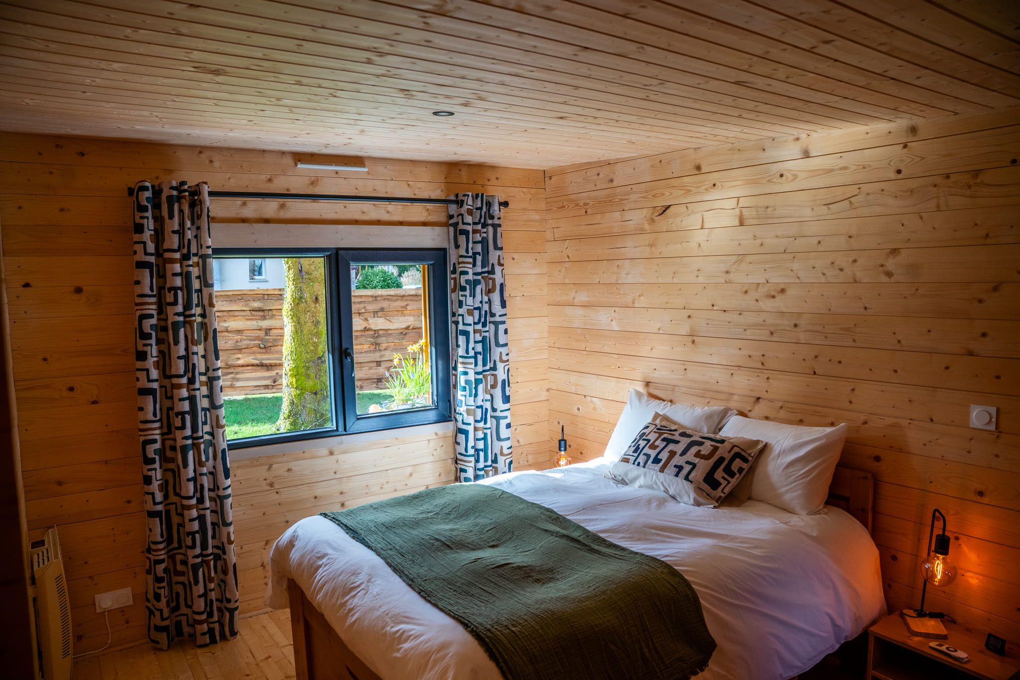 Un dormitorio de cabaña de madera con cama, cortinas, una ventana y una lámpara de noche.