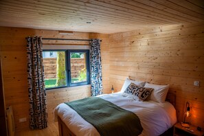 Un dormitorio de cabaña de madera con cama, cortinas, una ventana y una lámpara de noche.