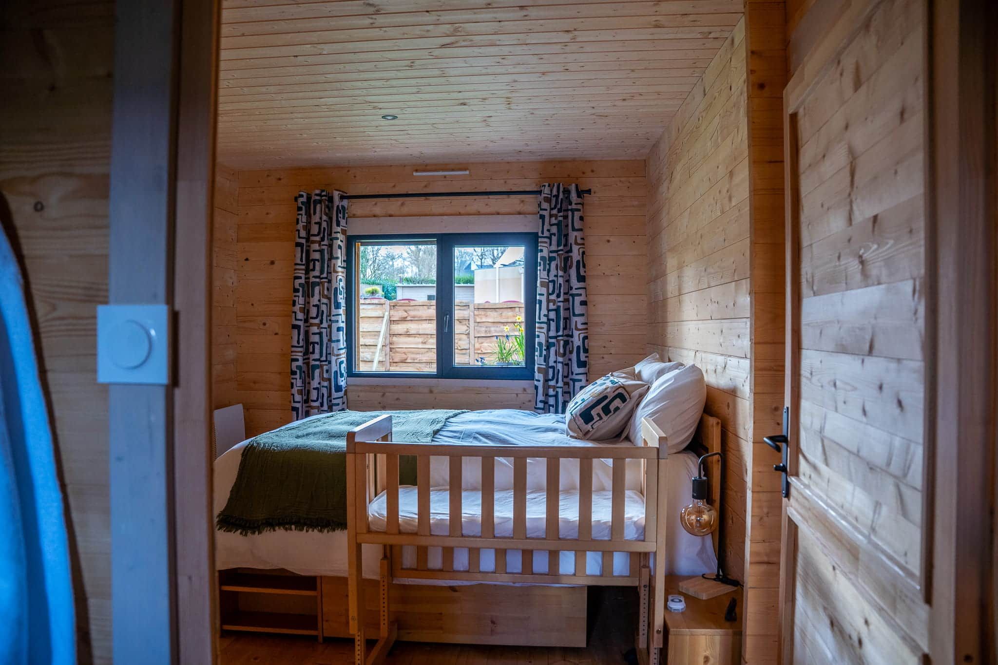 Ein kleines Zimmer mit Holzwänden, ausgestattet mit einem Etagenbett, einem Fenster mit Vorhängen und einer Lampe.