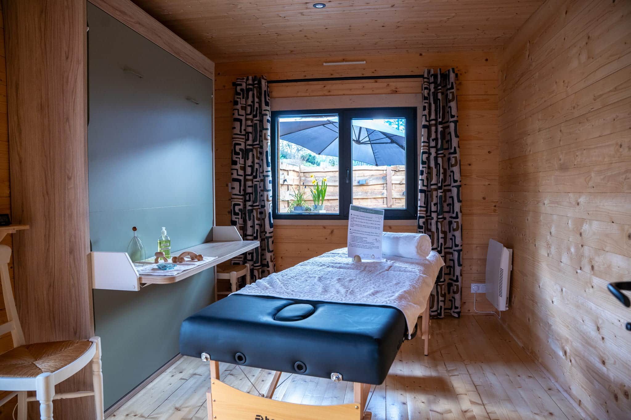 A room with a bed, a desk, and a chair. There is a window with curtains and a view of the outside.