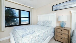 Ein Schlafzimmer mit einem Bett, einem Nachttisch, einer Lampe und einem Fenster mit Blick auf den Strand.