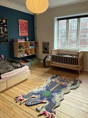 A room with a crib, a bookshelf, a large rug, and a window with a view of buildings. - Light and spacious flat in Nørrebro with garden (København)