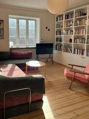 A cozy living room with a sofa, a pink chair, a bookshelf, and a large window. - Light and spacious flat in Nørrebro with garden (København)