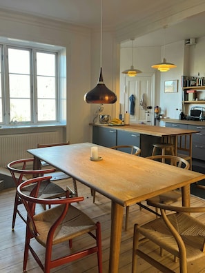 A kitchen with a wooden table, red chairs, a hanging pendant light, and a window with blinds. - Light and spacious flat in Nørrebro with garden (København)