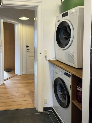 A laundry area with two washing machines, a wooden floor, and a door leading to another room. - Light and spacious flat in Nørrebro with garden (København)