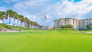 A golf course with a flagstick, palm trees, and a hotel building in the background. - 2206 Harbour Village (Ponce Inlet)