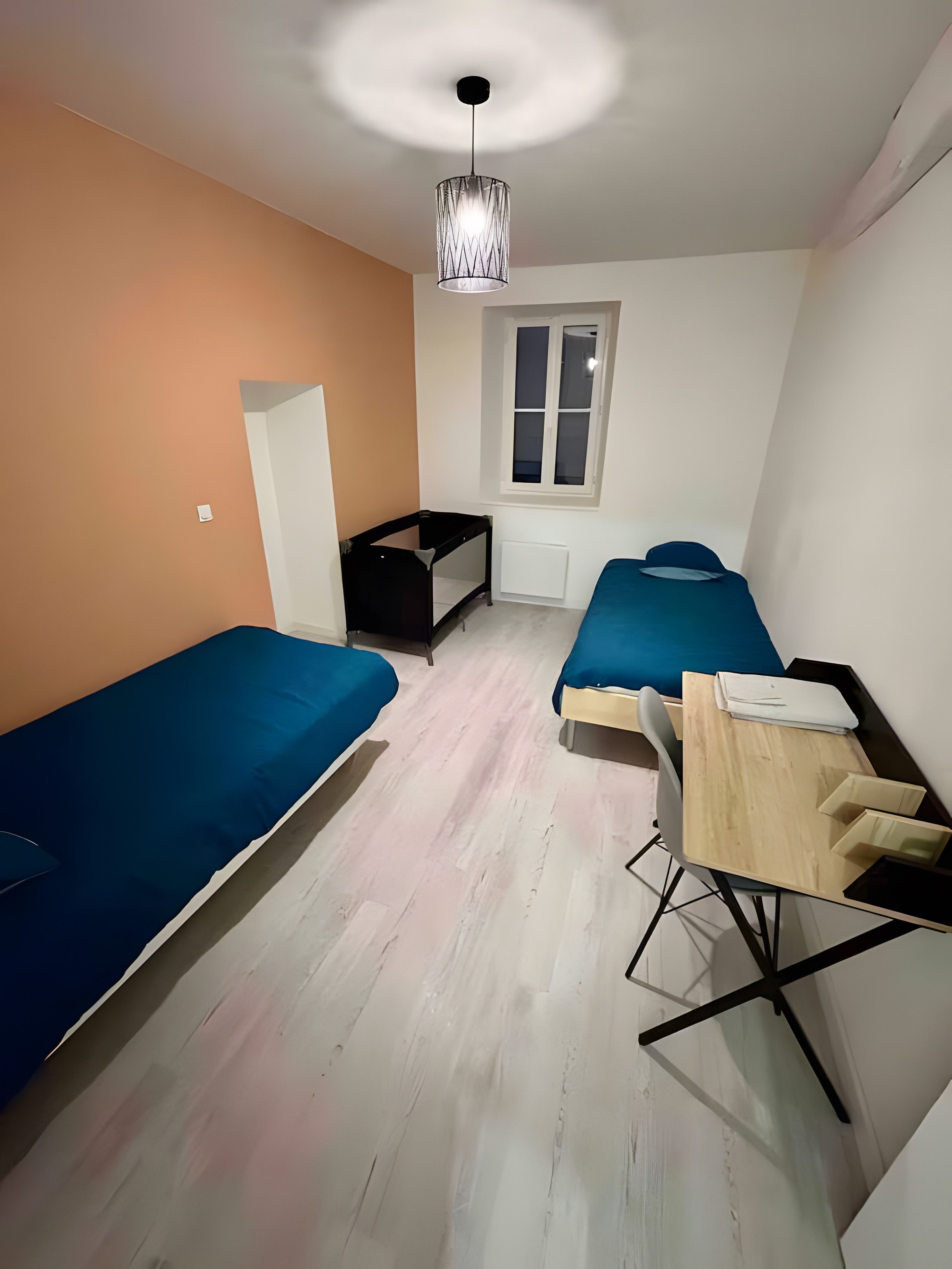 Une petite pièce, de taille modeste, comprenant deux lits simples, un bureau avec une chaise, un radiateur et une fenêtre.