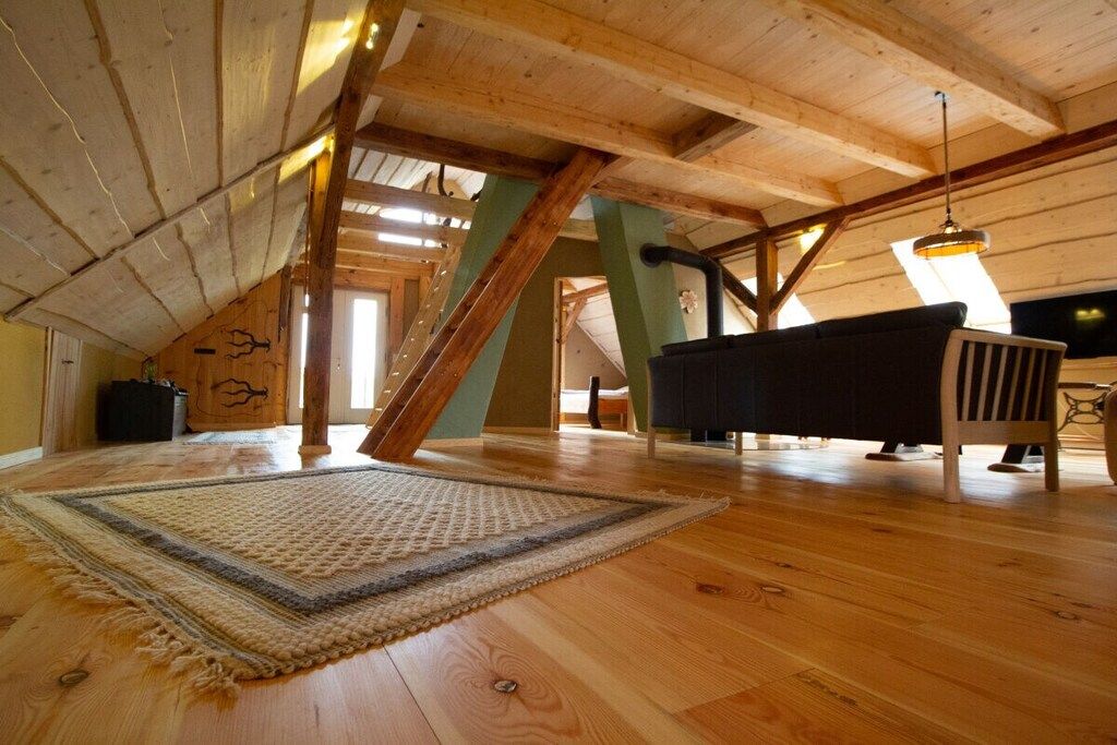 A spacious attic room with wooden beams, a sofa, a rug, and a hanging light fixture.