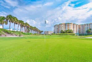 A golf course with a flagstick, palm trees, and residential buildings in the background. - 2306 Harbour Village (Ponce Inlet)